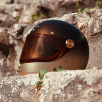 Full-face motorcycle helmet with a brown and beige gradient paint finish, placed on a rocky surface.